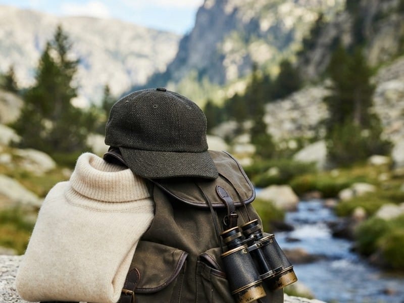 A folded cream turtleneck sweater, a dark cap, and vintage binoculars rest on a canvas backpack with a mountain stream in the background.