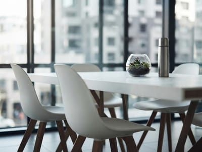 A white table and white modern chairs in an office with large windows overlooking city buildings, featuring a small glass succulent terrarium and a metal thermos on the tabletop.